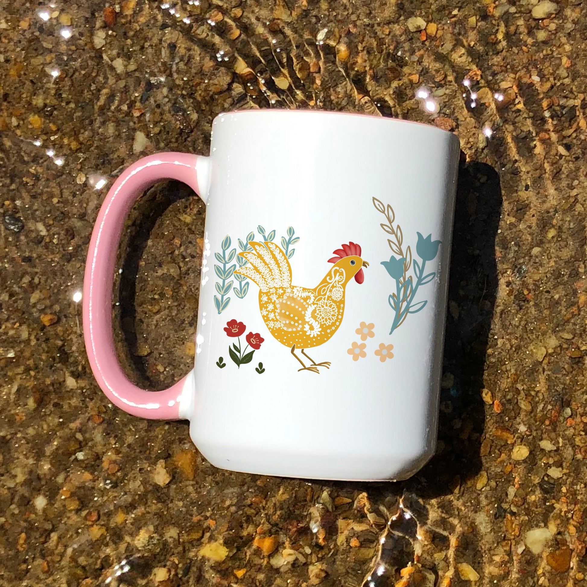 Chicken and rooster themed pink handle ceramic mug with floral designs in blue, yellow, and red with water ripples in the background.