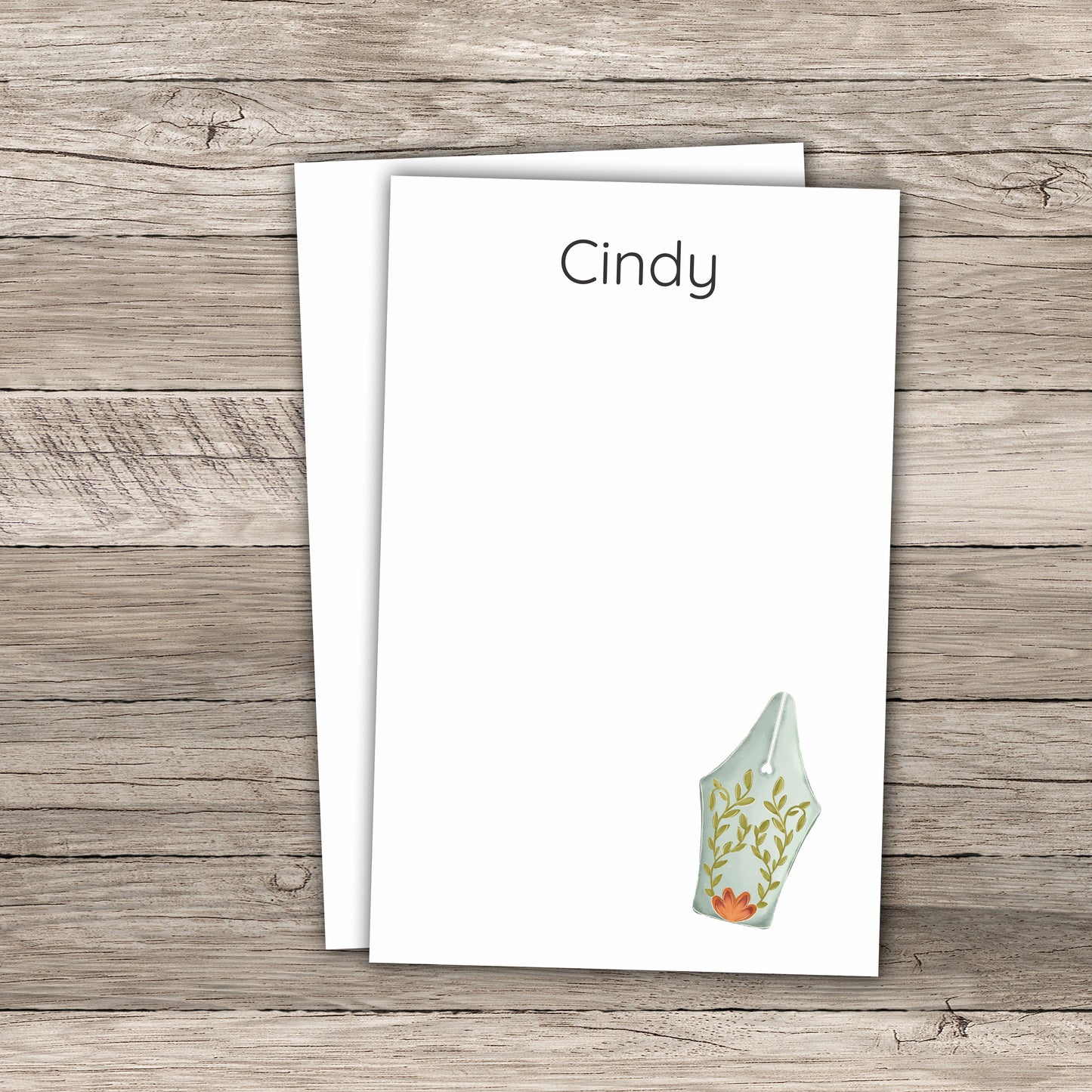Personalized notepad set featuring the name Cindy in block font at the top and a floral fountain pen illustration, printed on a white background with a wood-textured backdrop.