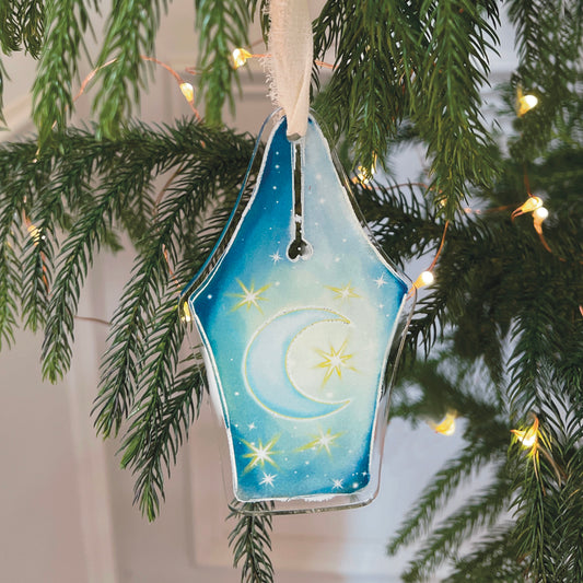 Acrylic fountain pen nib design ornament featuring moon and stars hanging against Christmas tree with lights.