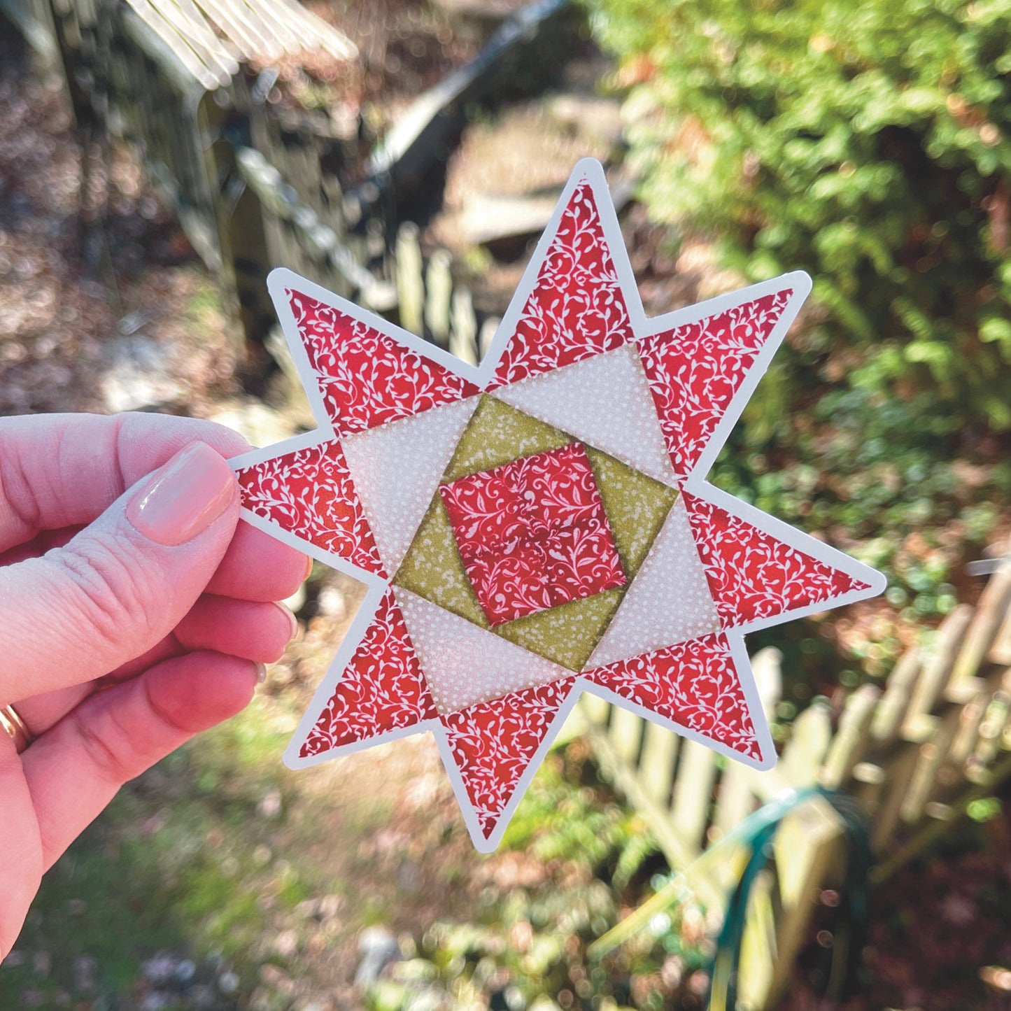 Medium Red, Green and Tan Quilt block sticker held over grassy outside background.