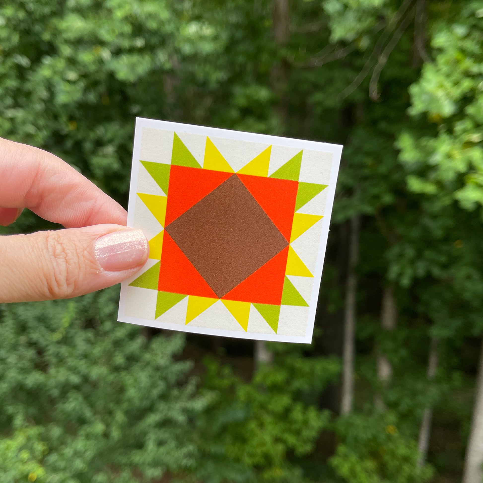 A hand holding a orange, yellow, and green barn quilt style square sticker against a blurred green background of trees.