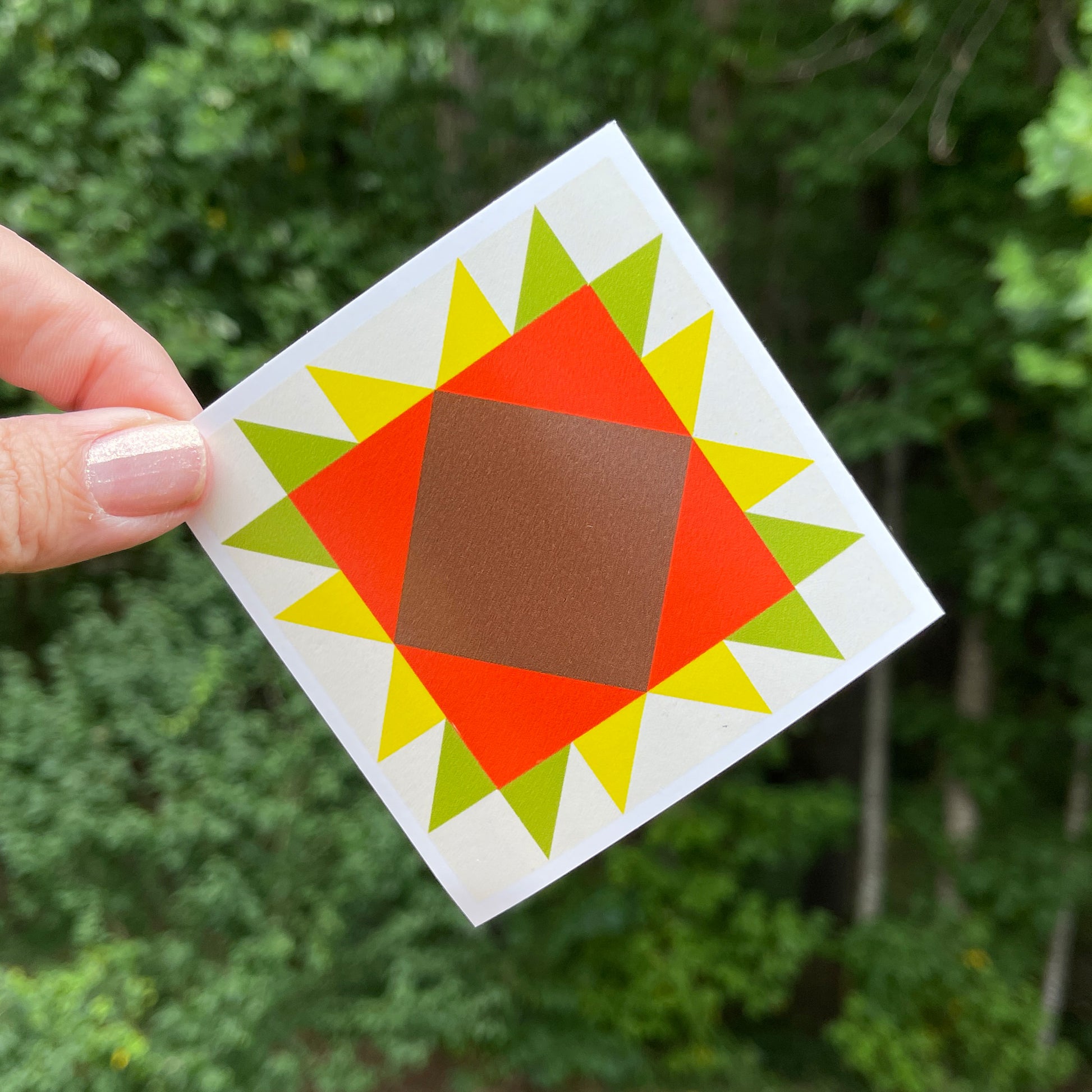 A hand holding a orange, yellow, and green barn quilt style square sticker against a blurred green background of trees.