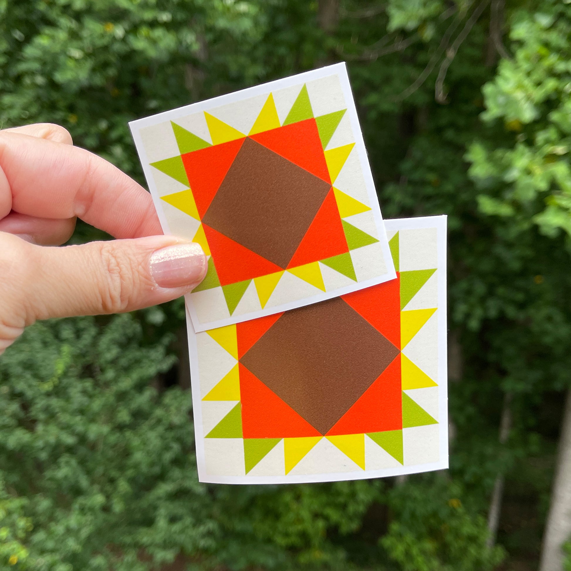 A hand holding two orange, yellow, and green barn quilt style square stickers against a blurred green background of trees.