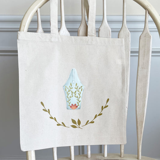 Natural cotton tote bag hanging on a white wooden chair, featuring a minimalist design of a fountain pen nib adorned with green leafy vines and a small orange flower. Below the pen nib, two curved branches form a simple, nature-inspired accent. 