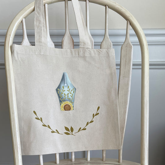 Natural canvas tote bag hanging on a wooden chair, featuring a decorative design of a fountain pen nib with sunflower and leaf motifs, and a delicate laurel wreath below.