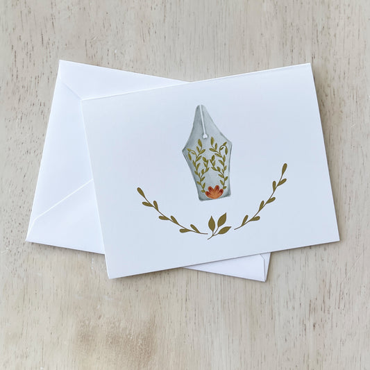 Hand-illustrated notecard featuring a fountain pen nib adorned with delicate floral and vine details, surrounded by simple botanical accents on a white background. The card is displayed with a matching white envelope on a light wooden surface.