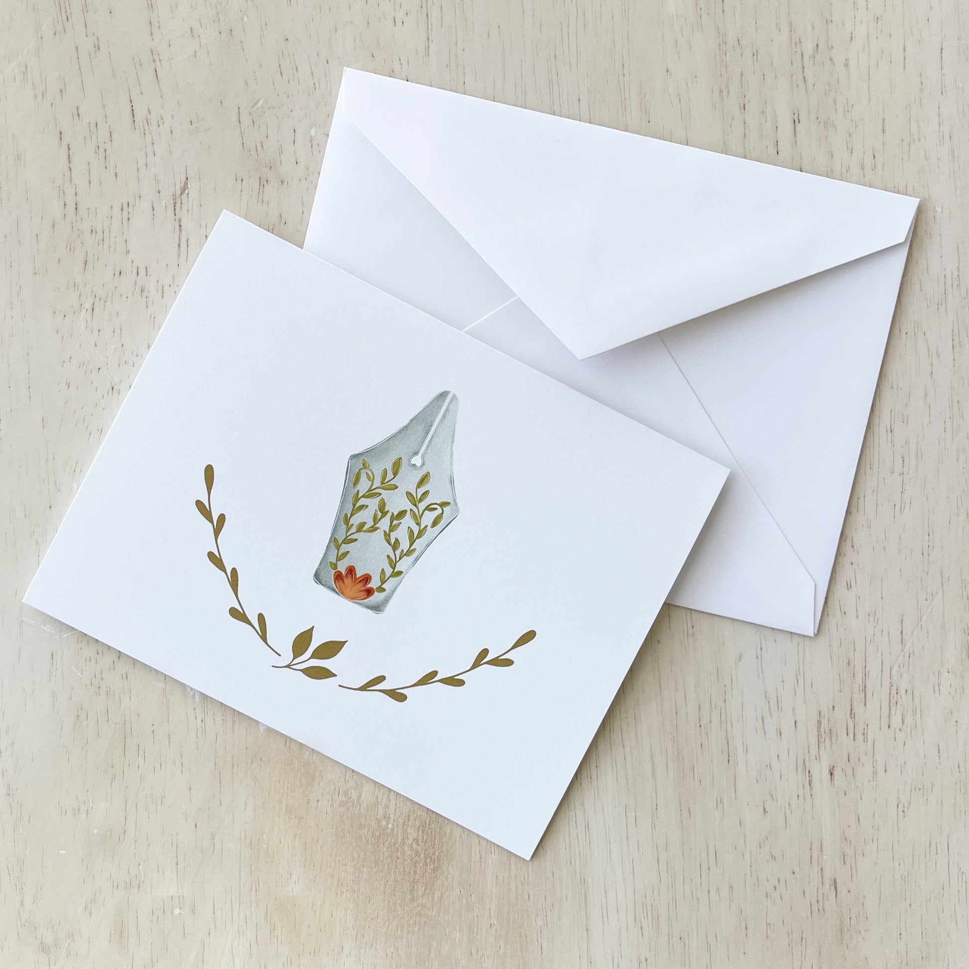 Hand-illustrated notecard featuring a fountain pen nib adorned with delicate floral and vine details, surrounded by simple botanical accents on a white background. The card is displayed with a matching white envelope on a light wooden surface.