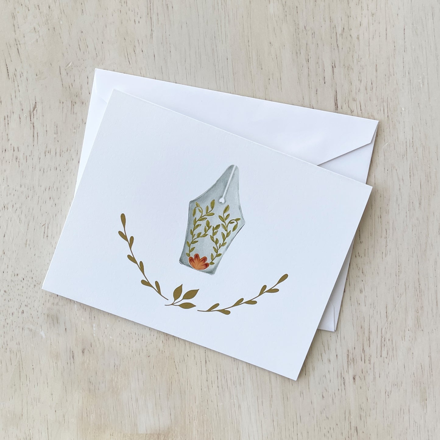 Hand-illustrated notecard featuring a fountain pen nib adorned with delicate floral and vine details, surrounded by simple botanical accents on a white background. The card is displayed with a matching white envelope on a light wooden surface.