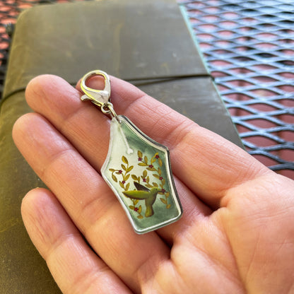 Close-up of a hand holding a hummingbird fountain pen nib acrylic charm with a lobster claw clasp. The charm features a detailed illustration of a hummingbird with leafy branches and small red flowers. A vintage-style journal is in the background.