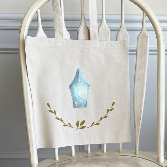 Natural cotton tote bag hanging on a white wooden chair, featuring a design of a fountain pen nib. The pen nib is illustrated with a crescent moon and stars against a soft blue background, with leafy branches curving upwards below the design.