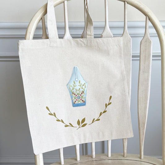 Natural cotton tote bag hanging on a white wooden chair, featuring a design of a fountain pen nib with a patchwork quilt block pattern and leafy vine accents. Below the pen nib, two green leafy branches curve upward, adding a decorative touch.