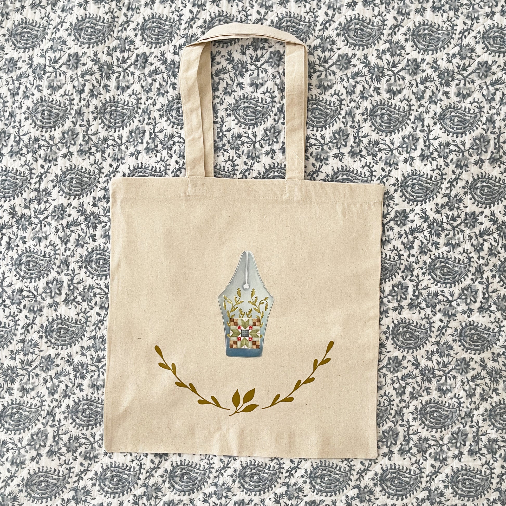 Natural cotton tote bag laying flat on paisley background, featuring a design of a fountain pen nib with a patchwork quilt block pattern and leafy vine accents. Below the pen nib, two green leafy branches curve upward, adding a decorative touch.