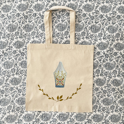 Natural cotton tote bag laying flat on paisley background, featuring a design of a fountain pen nib with a patchwork quilt block pattern and leafy vine accents. Below the pen nib, two green leafy branches curve upward, adding a decorative touch.