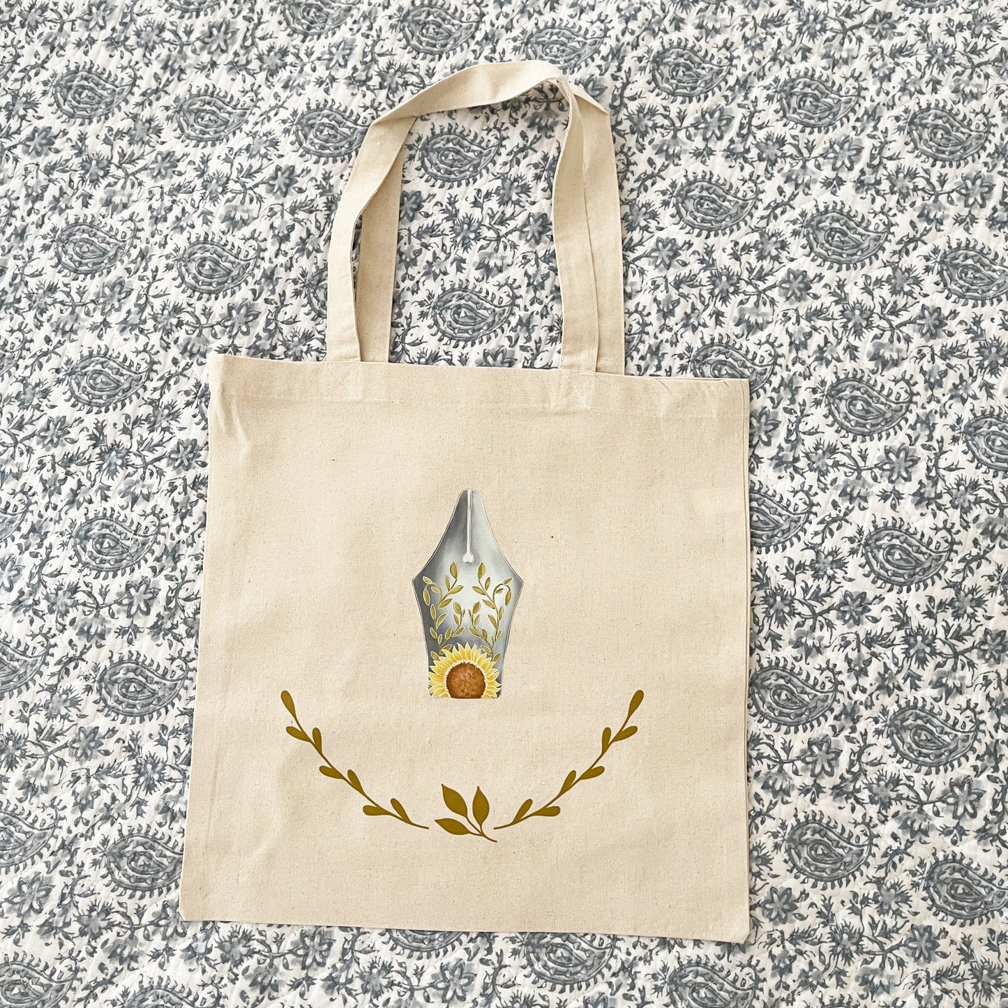 Natural canvas tote bag laying on paisley quilt, featuring a decorative design of a fountain pen nib with sunflower and leaf motifs, and a delicate laurel wreath below.