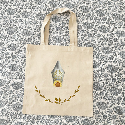 Natural canvas tote bag laying on paisley quilt, featuring a decorative design of a fountain pen nib with sunflower and leaf motifs, and a delicate laurel wreath below.