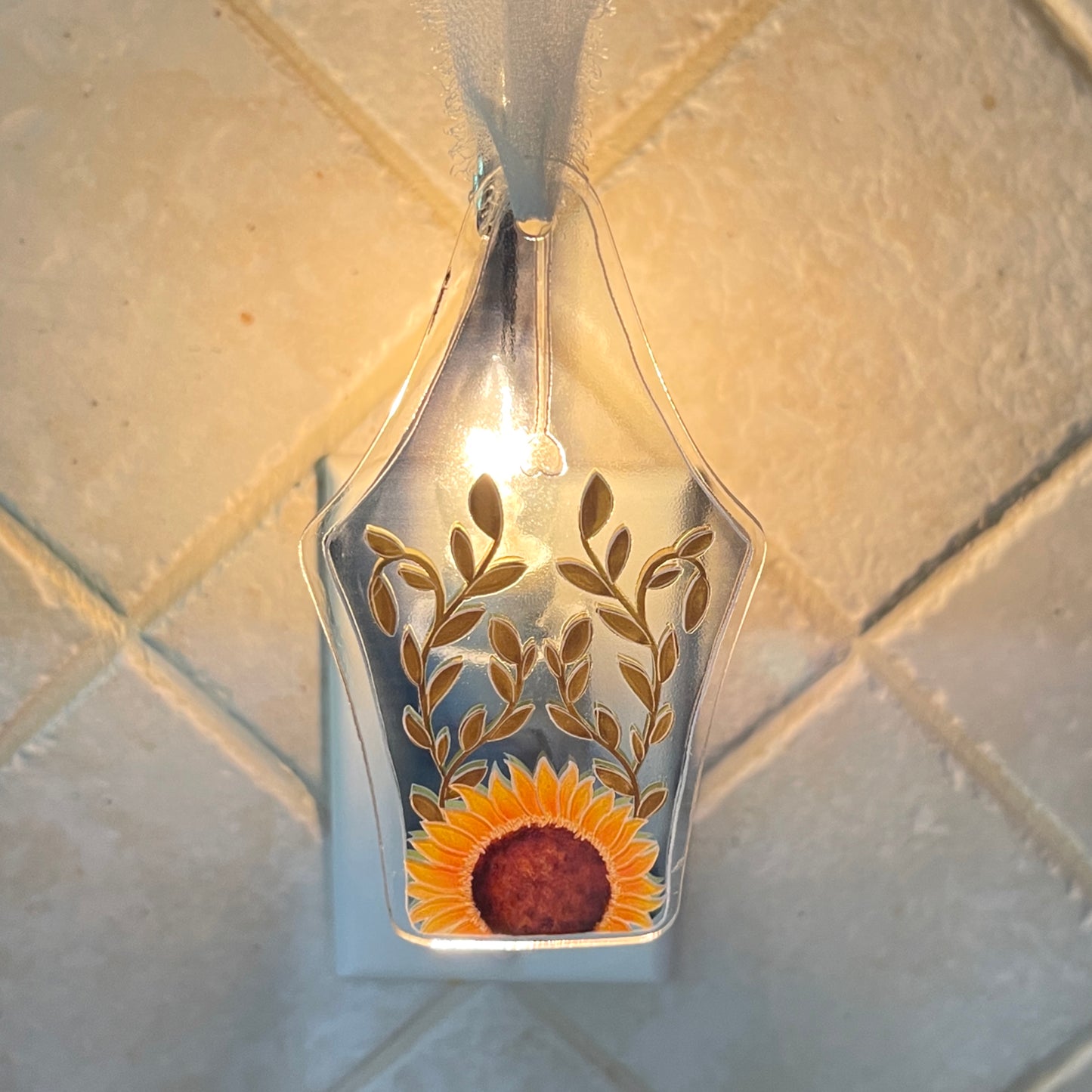 Clear acrylic ornament shaped like a fountain pen nib, featuring a sunflower surrounded by leaves. The ornament hanging by a white ribbon is illuminated by a light.