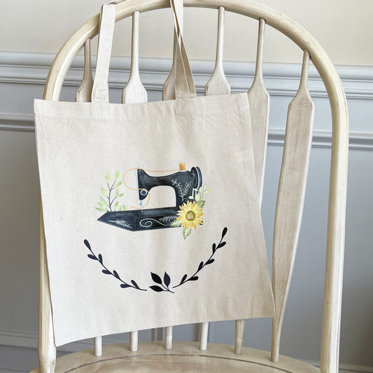 A beige canvas tote bag hanging on a wooden chair, featuring a hand-painted vintage sewing machine illustration adorned with green leaves and a sunflower. Below the sewing machine, black laurel leaves create a decorative accent.