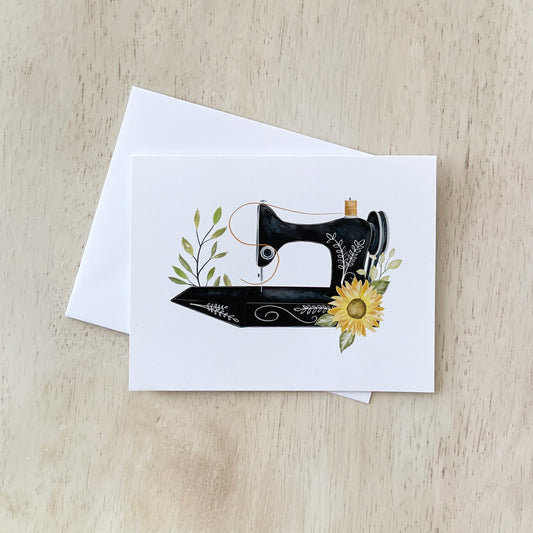 A greeting card featuring an illustration of a vintage black sewing machine adorned with delicate greenery and a bright yellow sunflower displayed on a light wooden surface, accompanied by a plain white envelope.