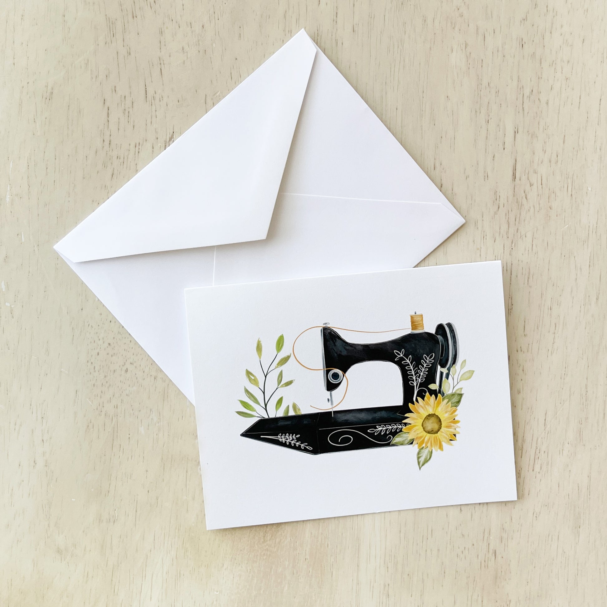 A greeting card featuring an illustration of a vintage black sewing machine adorned with delicate greenery and a bright yellow sunflower displayed on a light wooden surface, accompanied by a plain white envelope.