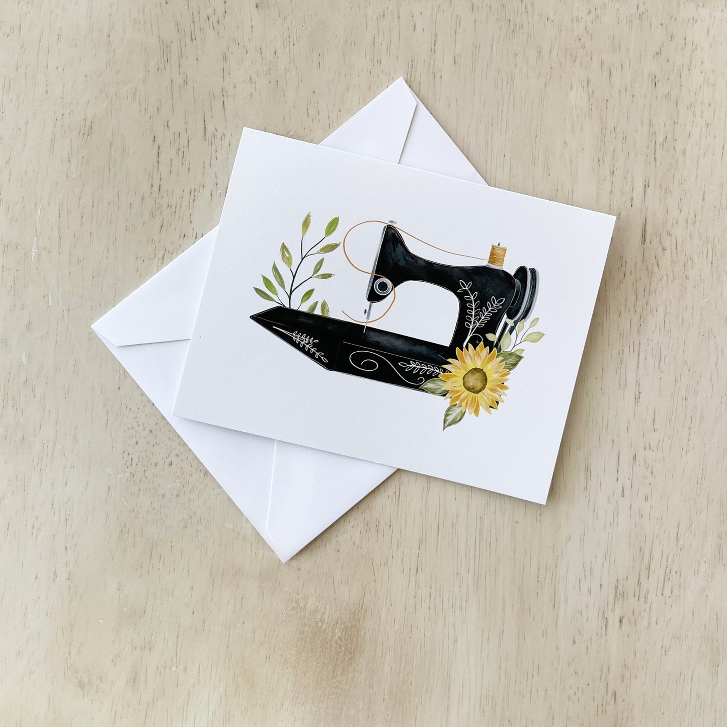 A greeting card featuring an illustration of a vintage black sewing machine adorned with delicate greenery and a bright yellow sunflower displayed on a light wooden surface, accompanied by a plain white envelope.