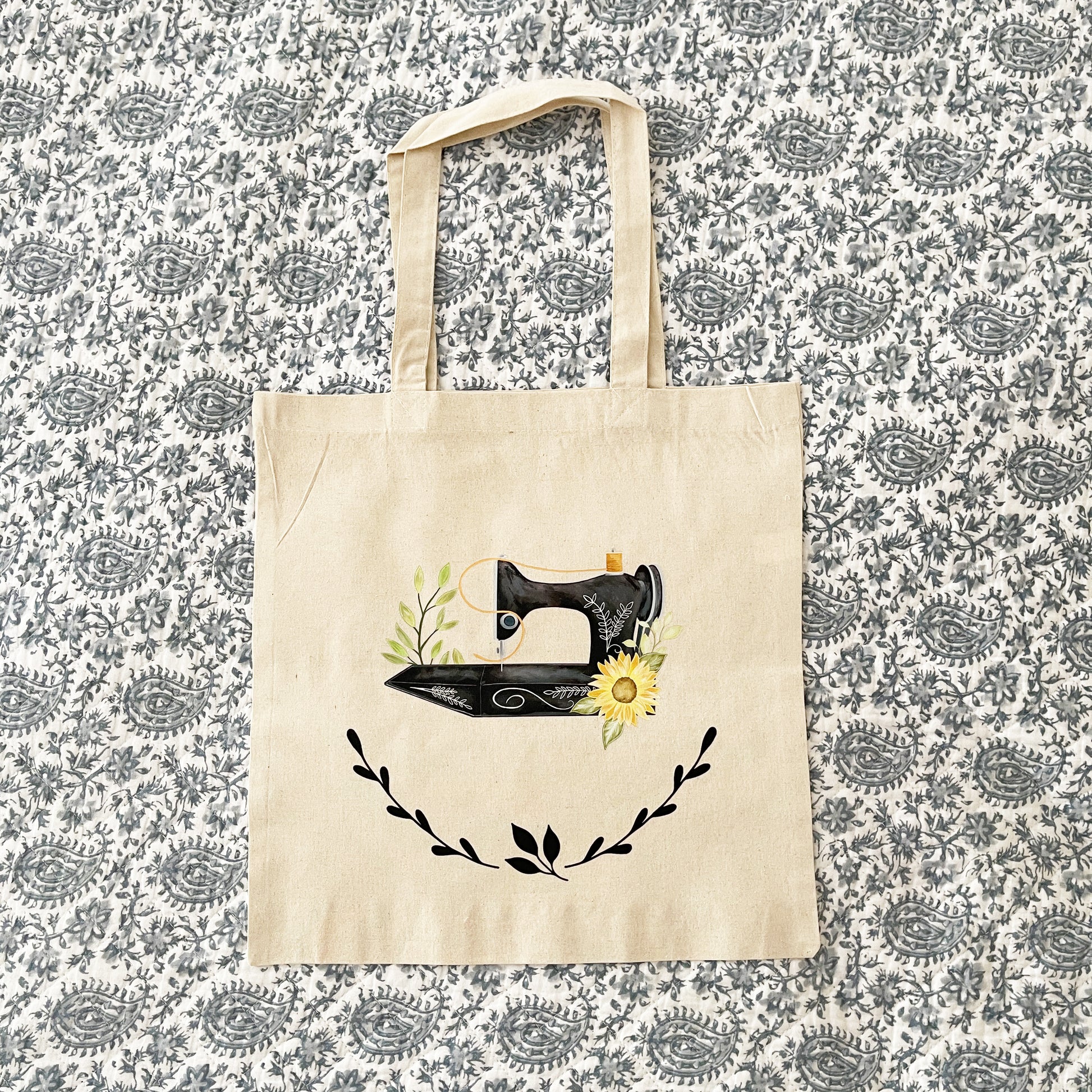 A beige canvas tote bag laying on a paisley pattern quilt, featuring a hand-painted vintage sewing machine illustration adorned with green leaves and a sunflower. Below the sewing machine, black laurel leaves create a decorative accent.