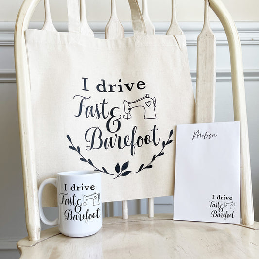 Set of sewing-themed gifts featuring a natural canvas tote bag, white ceramic mug, and personalized notepad, all printed with the phrase 'I drive Fast & Barefoot' with a simple sewing machine illustration. The items are displayed on a wooden chair.