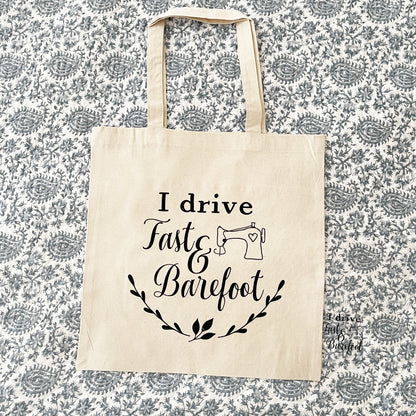 Natural cotton tote bag laying flat on blue paisley background, featuring the phrase 'I drive fast & barefoot' in black script with an illustration of a sewing machine and decorative leaf accents below. 