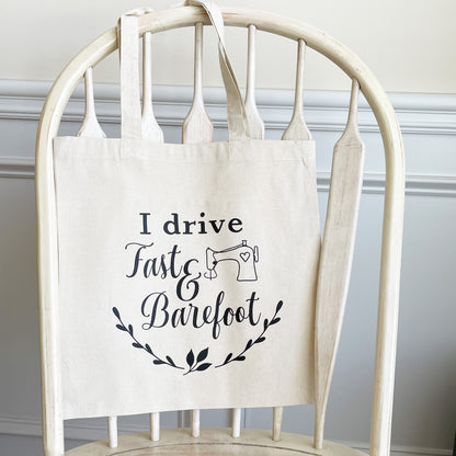 Natural cotton tote bag hanging on a white wooden chair, featuring the phrase 'I drive fast & barefoot' in black script with an illustration of a sewing machine and decorative leaf accents below. 