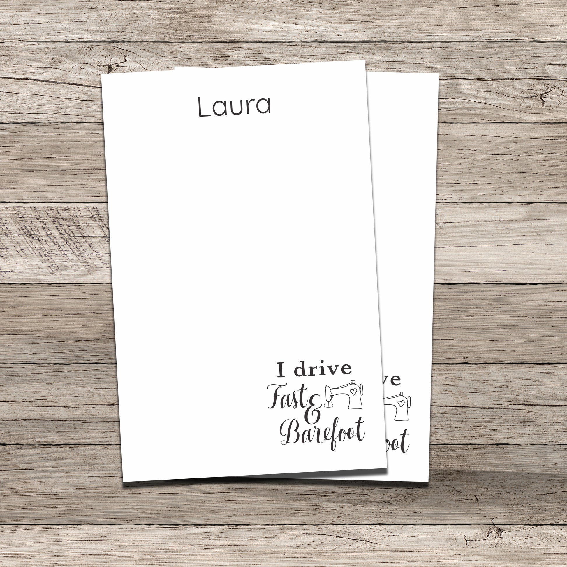 Personalized notepads featuring the name 'Laura' at the top and a sewing-themed phrase at the bottom that reads 'I drive Fast & Barefoot' with a sewing machine illustration, printed on a white background with a wood-textured backdrop.