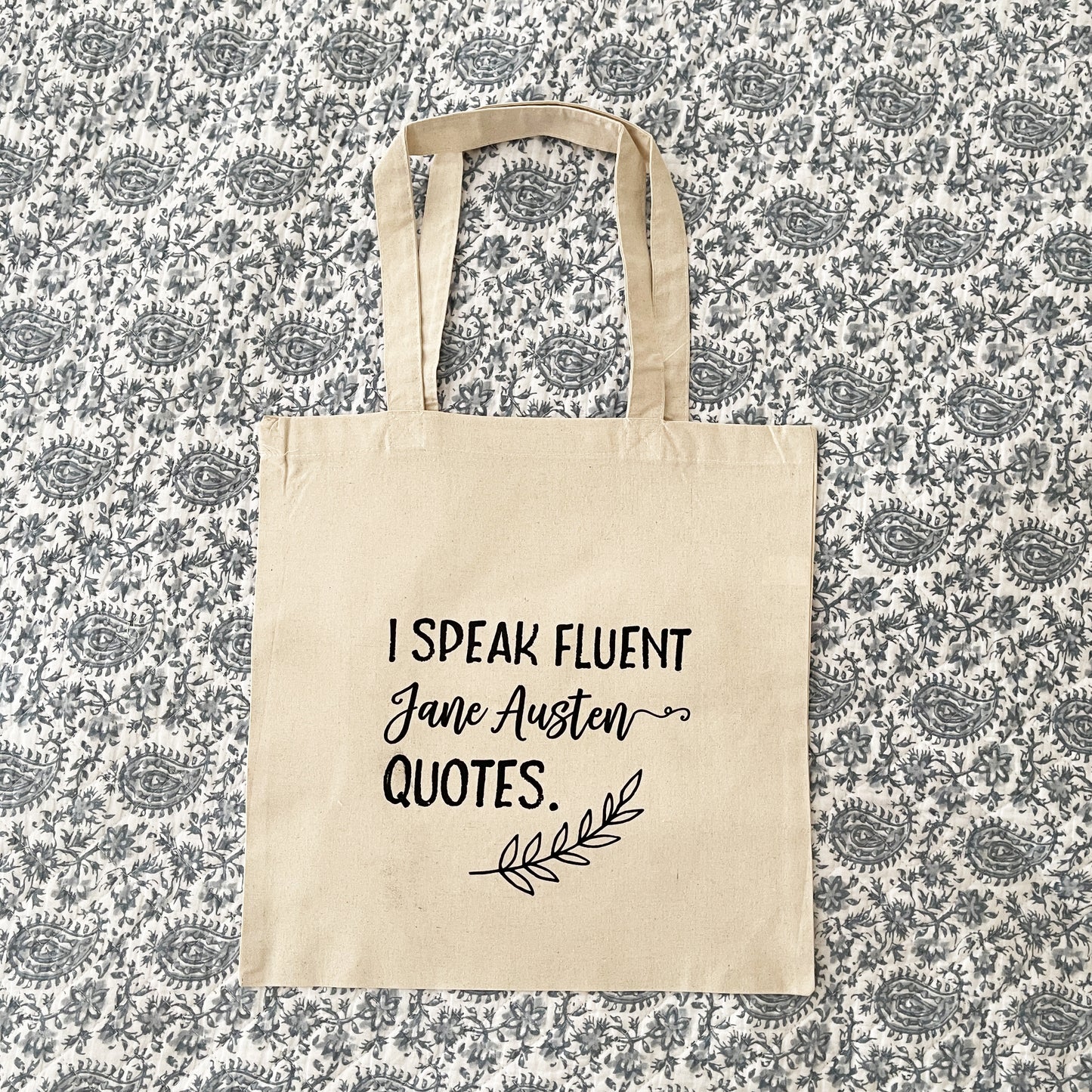 Natural cotton tote bag laying flat on a paisley background, featuring the phrase 'I speak fluent Jane Austen quotes.' in bold black script, with a decorative leaf accent below, perfect for book lovers and Austen fans.