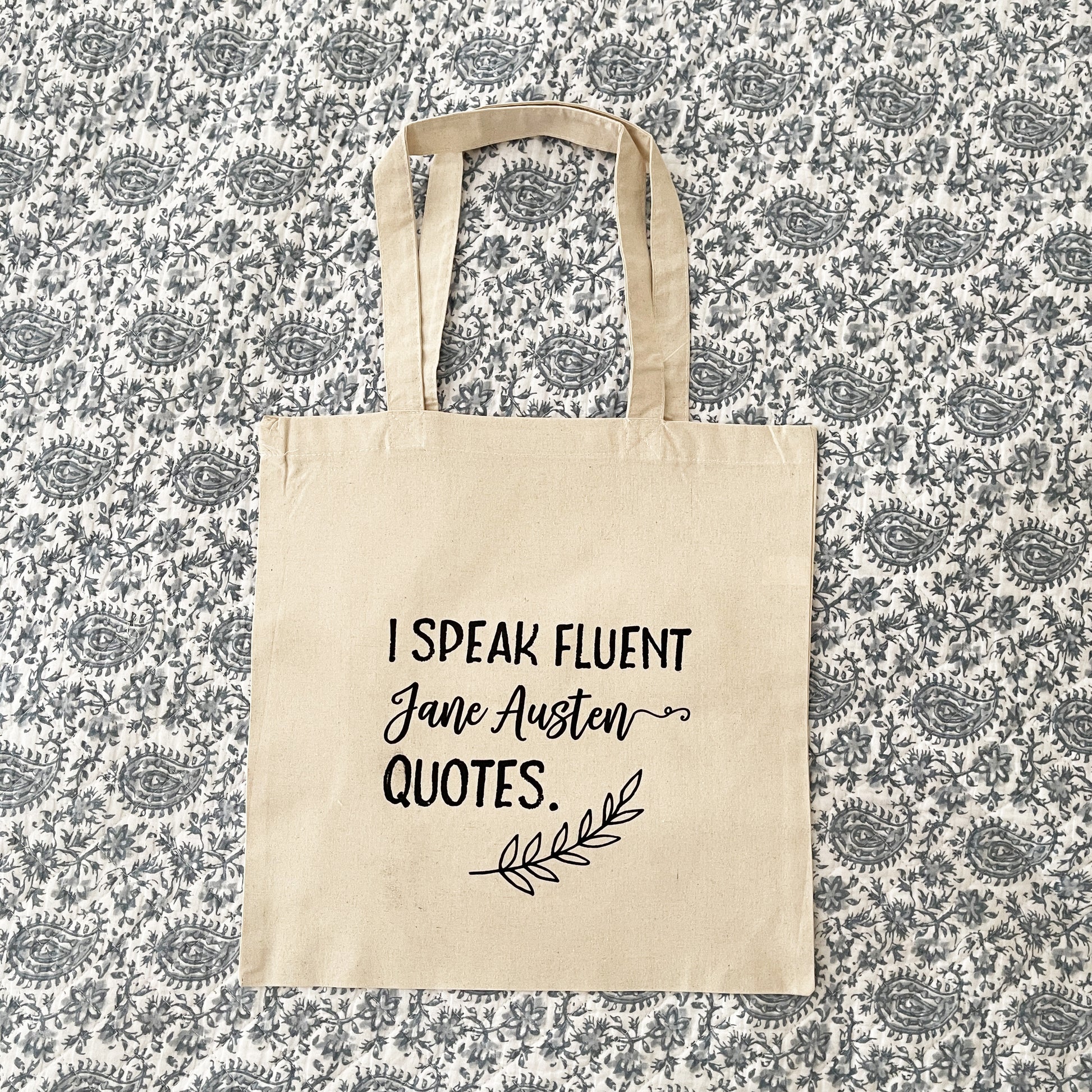 Natural cotton tote bag laying flat on a paisley background, featuring the phrase 'I speak fluent Jane Austen quotes.' in bold black script, with a decorative leaf accent below, perfect for book lovers and Austen fans.