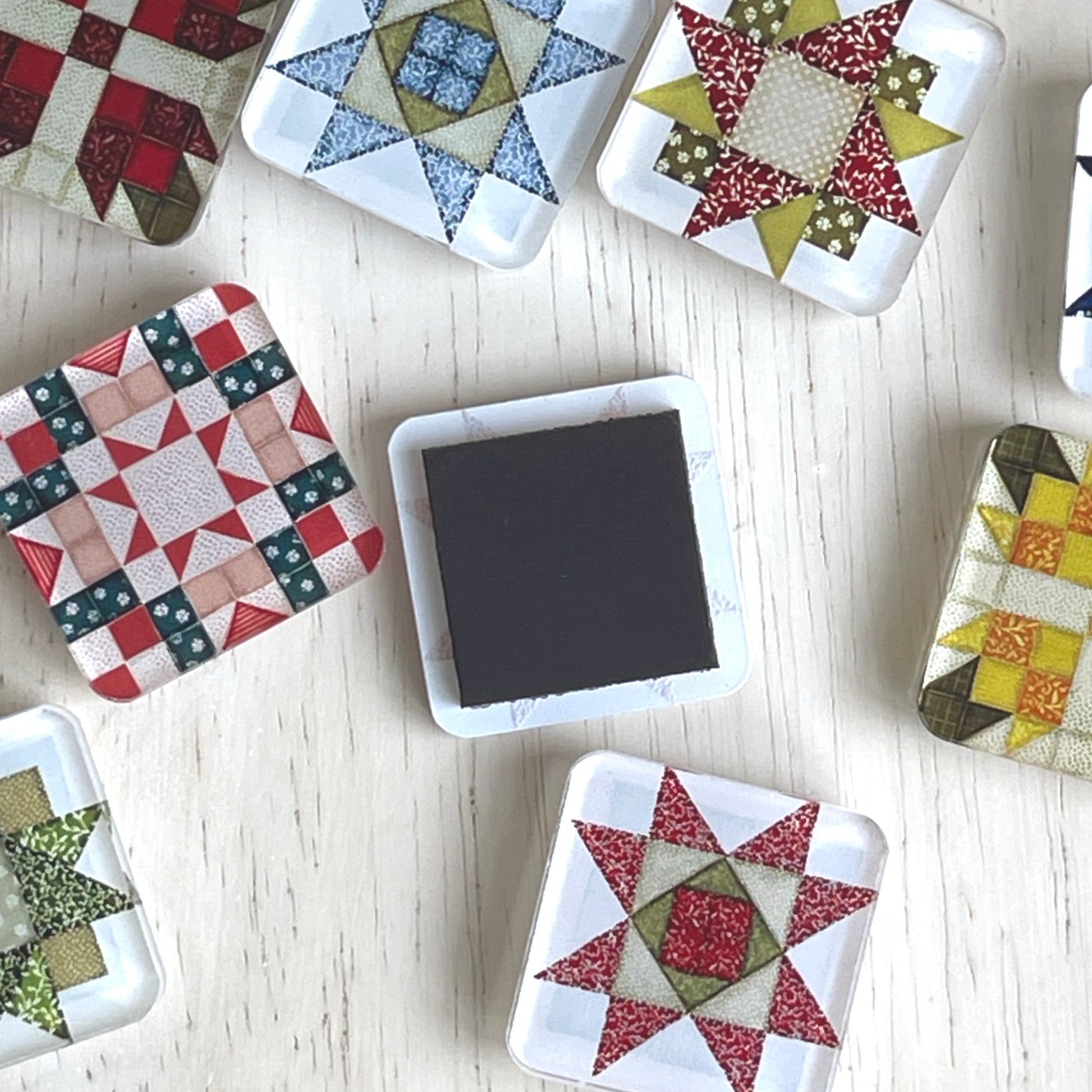 Scattered square magnets featuring various quilt block designs in different colors, including red, green, blue, and yellow. One magnet is flipped over to show the magnet backing. The magnets are arranged on a light wooden surface.