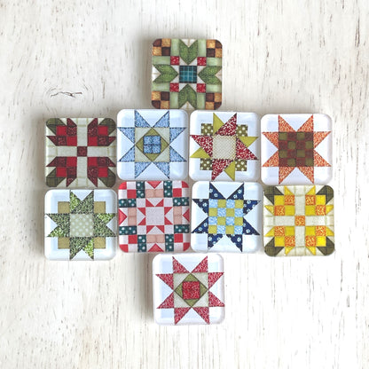 Set of ten square magnets featuring various quilt block designs in different colors, including red, green, blue, and yellow. The magnets are arranged on a light wooden surface, each showcasing a unique patchwork pattern.