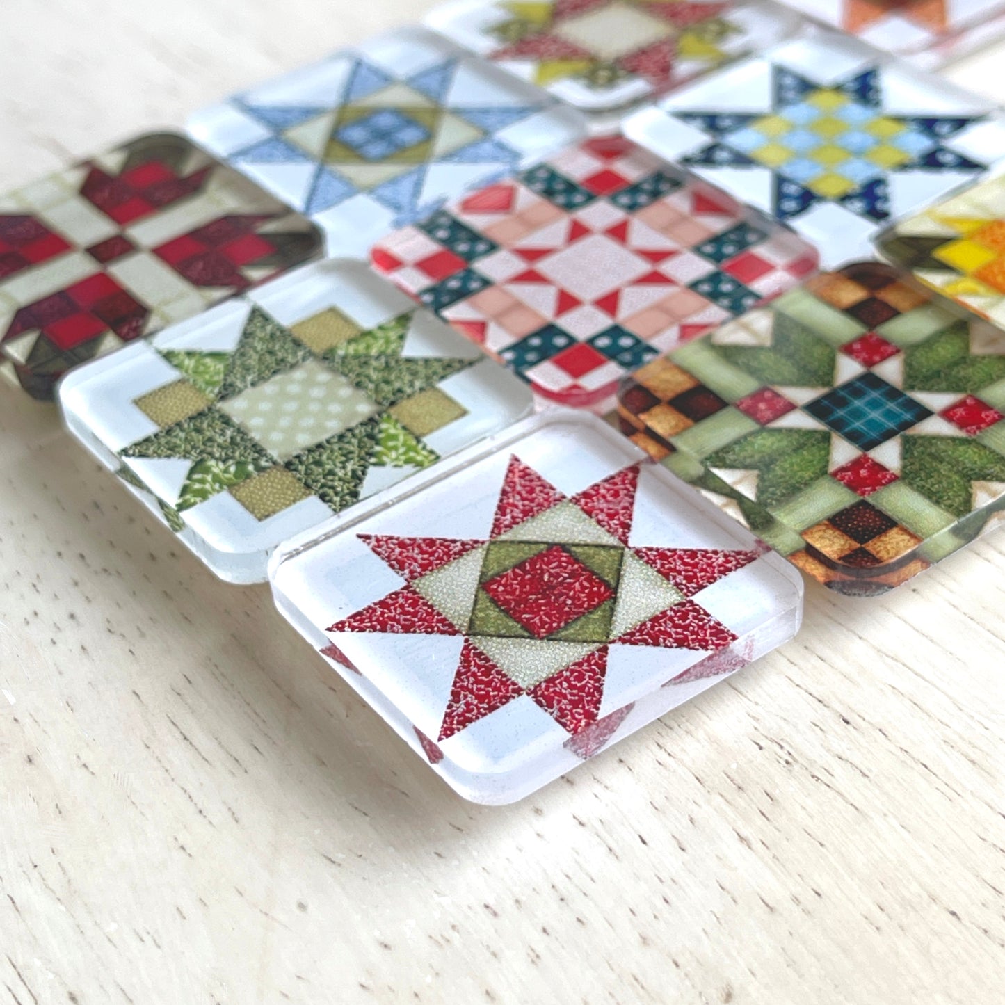 Side view of square magnets featuring various quilt block designs in different colors, including red, green, blue, and yellow. The magnets are arranged on a light wooden surface, each showcasing a unique patchwork pattern.