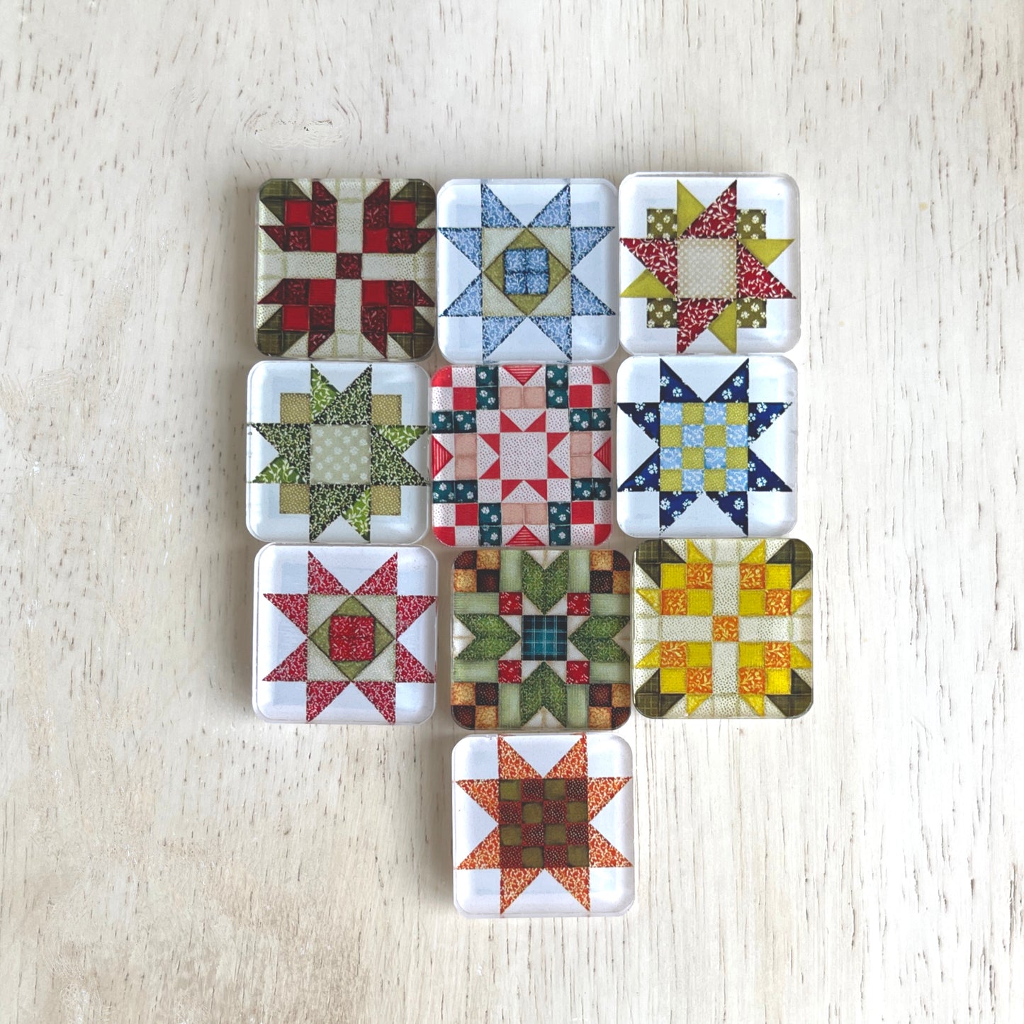 Set of ten square magnets featuring various quilt block designs in different colors, including red, green, blue, and yellow. The magnets are arranged on a light wooden surface, each showcasing a unique patchwork pattern.