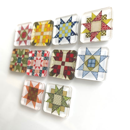 Set of ten square magnets featuring various quilt block designs in different colors, including red, green, blue, and yellow. The magnets are arranged on a white surface, each showcasing a unique patchwork pattern.