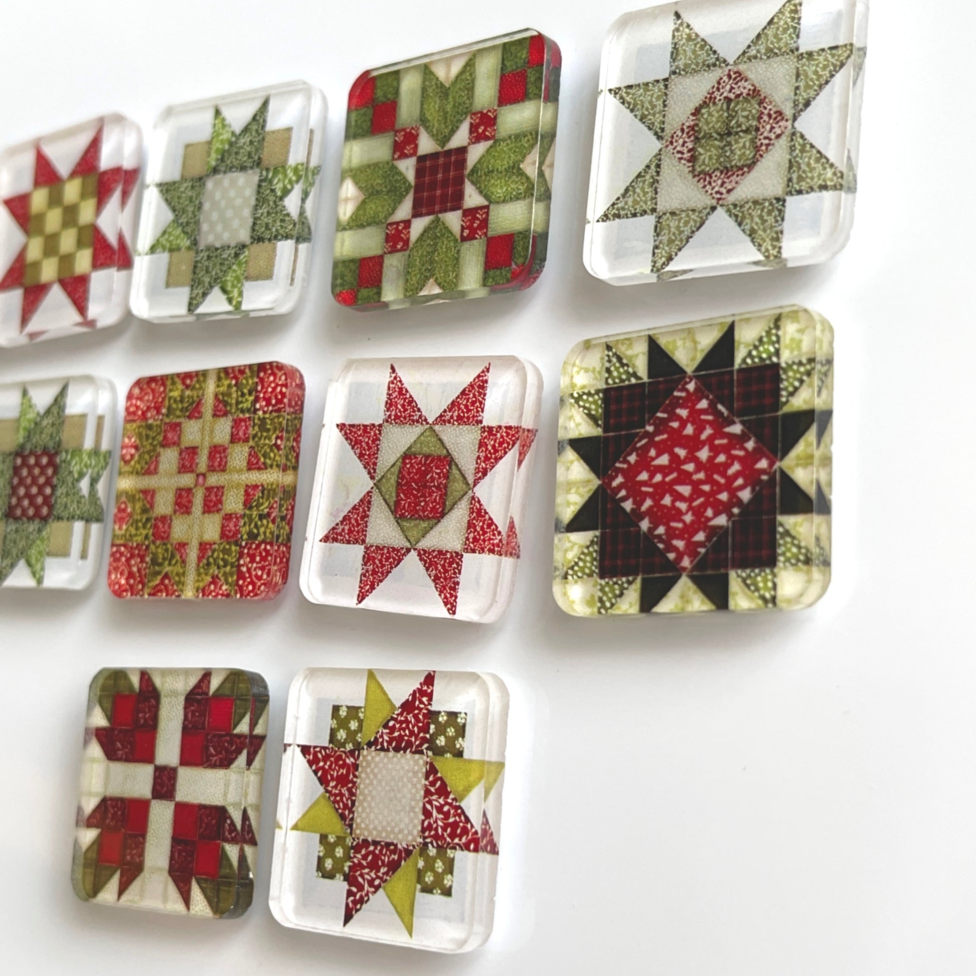 Side view of set of ten square magnets featuring colorful Christmas-themed quilt block designs in traditional red, green, and white patterns. Each magnet showcases a unique quilt patchwork design, arranged in a neat grid on a white background.