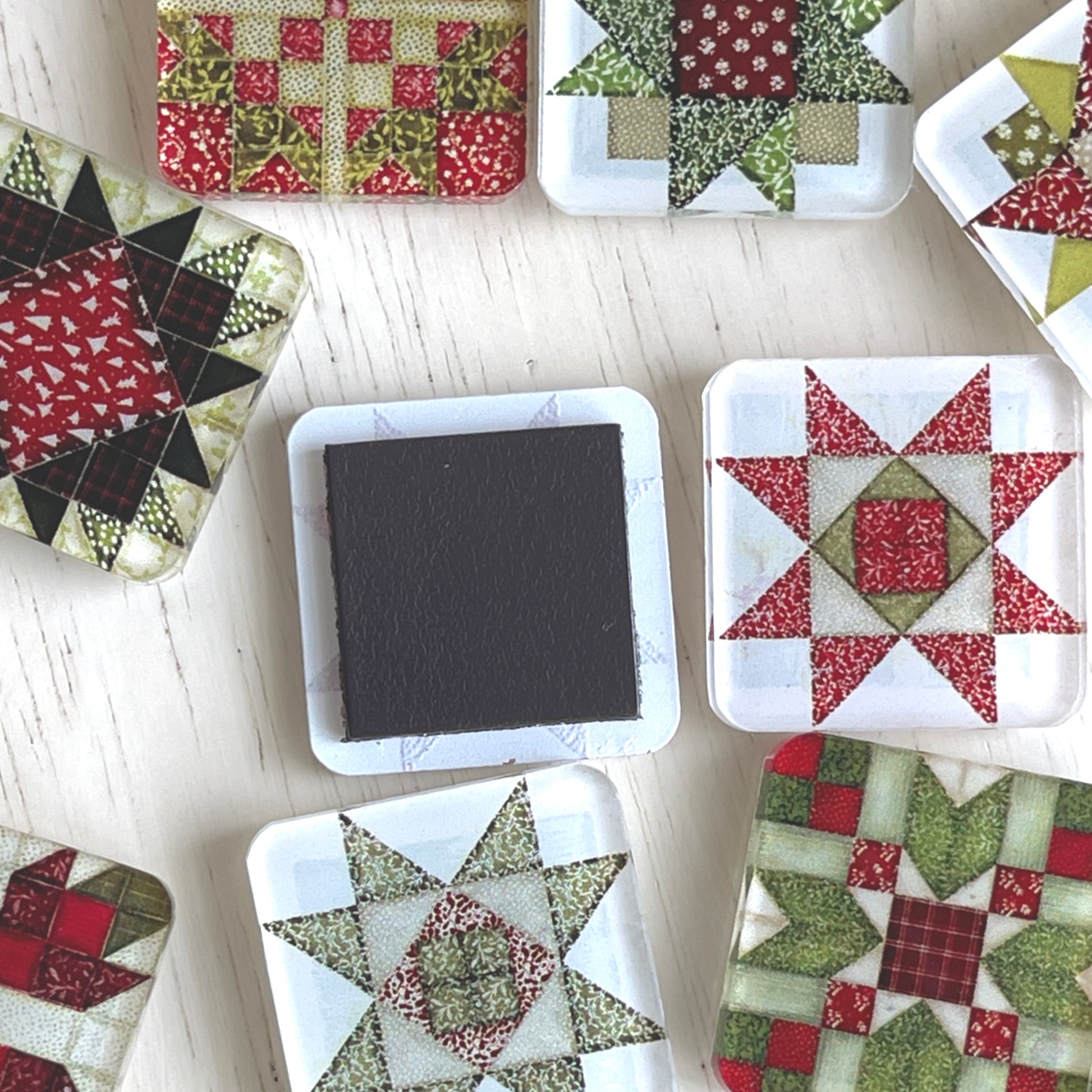 Scattered square magnets featuring colorful Christmas-themed quilt block designs in traditional red, green, and white patterns. One magnet is flipped to show the magnet on back.