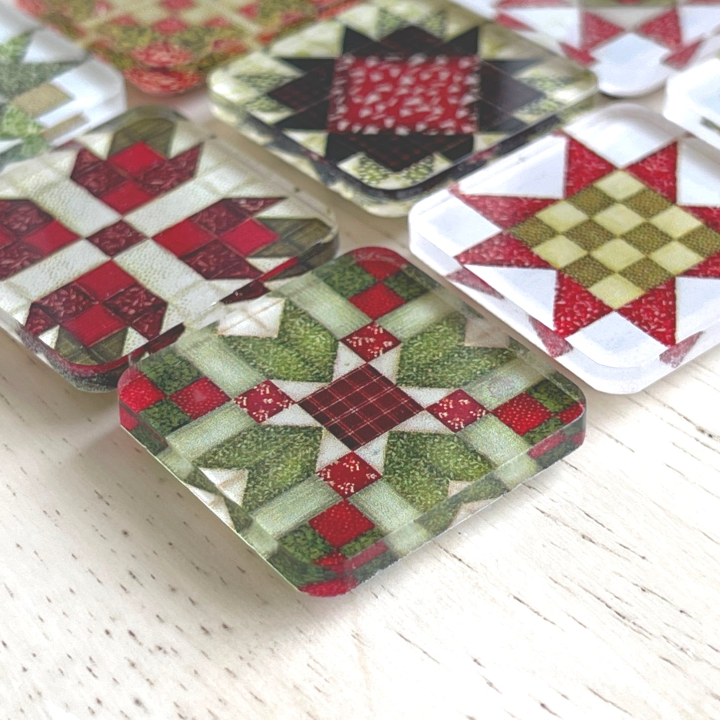 Close up square magnets featuring colorful Christmas-themed quilt block designs in traditional red, green, and white patterns. Each magnet showcases a unique quilt patchwork design, arranged in a neat grid on a weathered background.