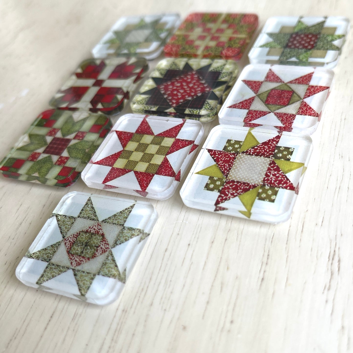 Set of ten square magnets featuring colorful Christmas-themed quilt block designs in traditional red, green, and white patterns. Each magnet showcases a unique quilt patchwork design, arranged in a neat grid on a weathered background.