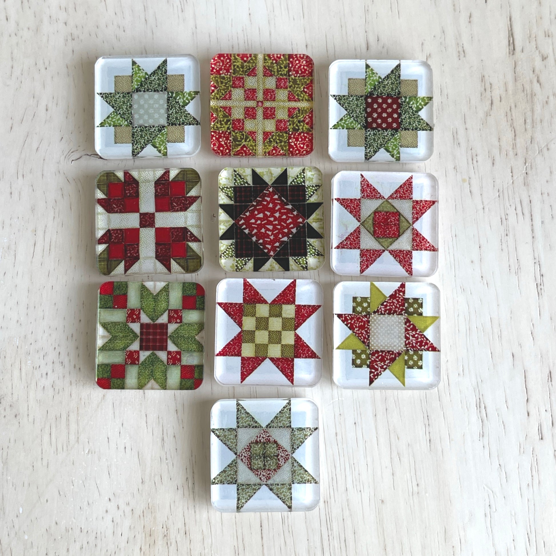 Set of ten square magnets featuring colorful Christmas-themed quilt block designs in traditional red, green, and white patterns. Each magnet showcases a unique quilt patchwork design, arranged in a neat grid on a weathered background.