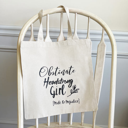 Natural cotton tote bag hanging on a white wooden chair, featuring the quote 'Obstinate Headstrong Girl' in bold black script, with 'Pride & Prejudice' written below in smaller text. The design also includes a small leafy accent, giving the tote a literary and minimalist aesthetic.