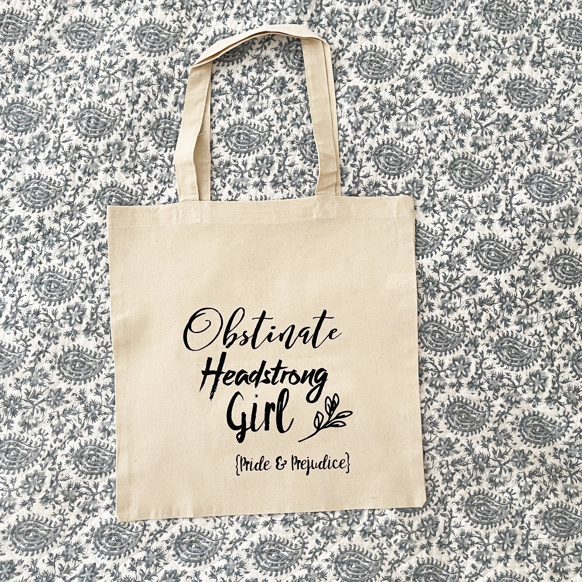 Natural cotton tote bag laying flat on paisley background, featuring the quote 'Obstinate Headstrong Girl' in bold black script, with 'Pride & Prejudice' written below in smaller text. The design also includes a small leafy accent.