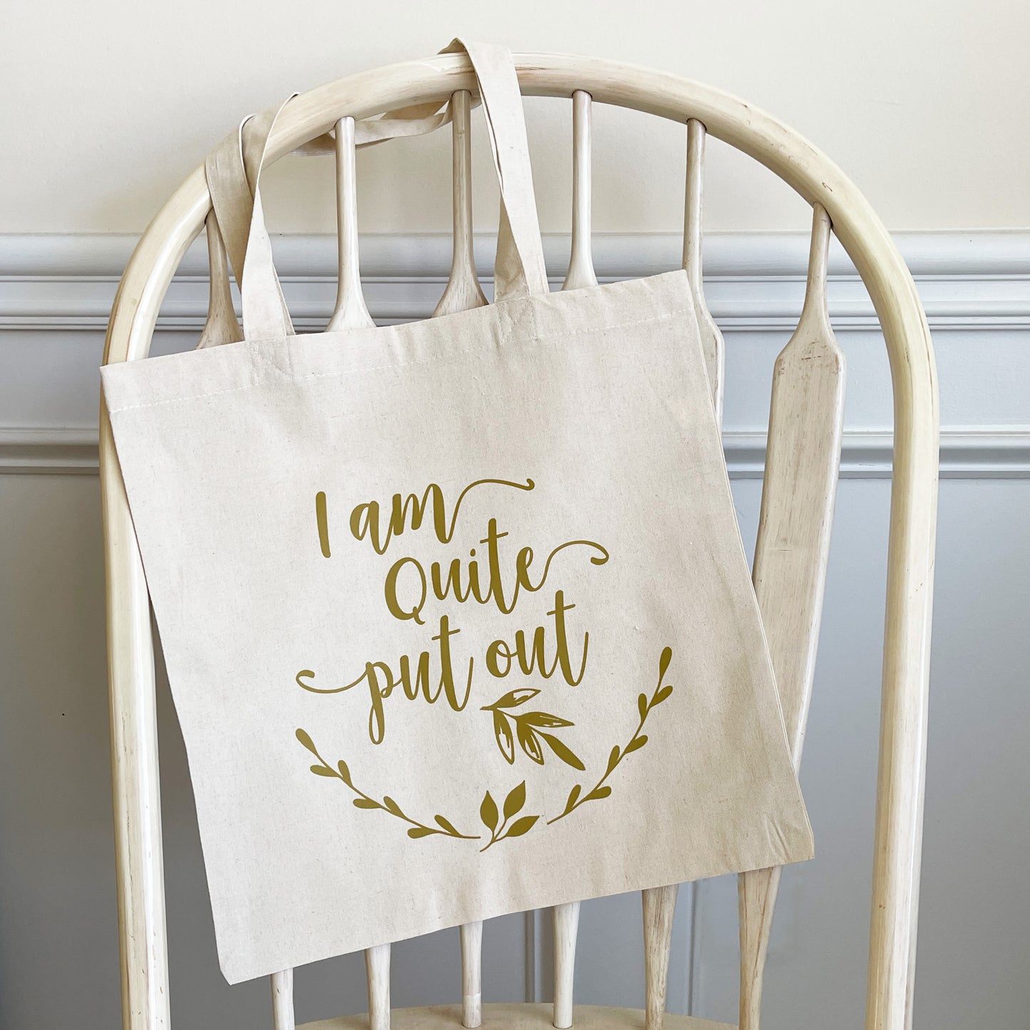 Natural cotton tote bag hanging on a white wooden chair, featuring the phrase 'I am quite put out' in elegant script with a leafy branch accent below. The text and design are printed in a soft, muted color, creating a minimalist and playful aesthetic.