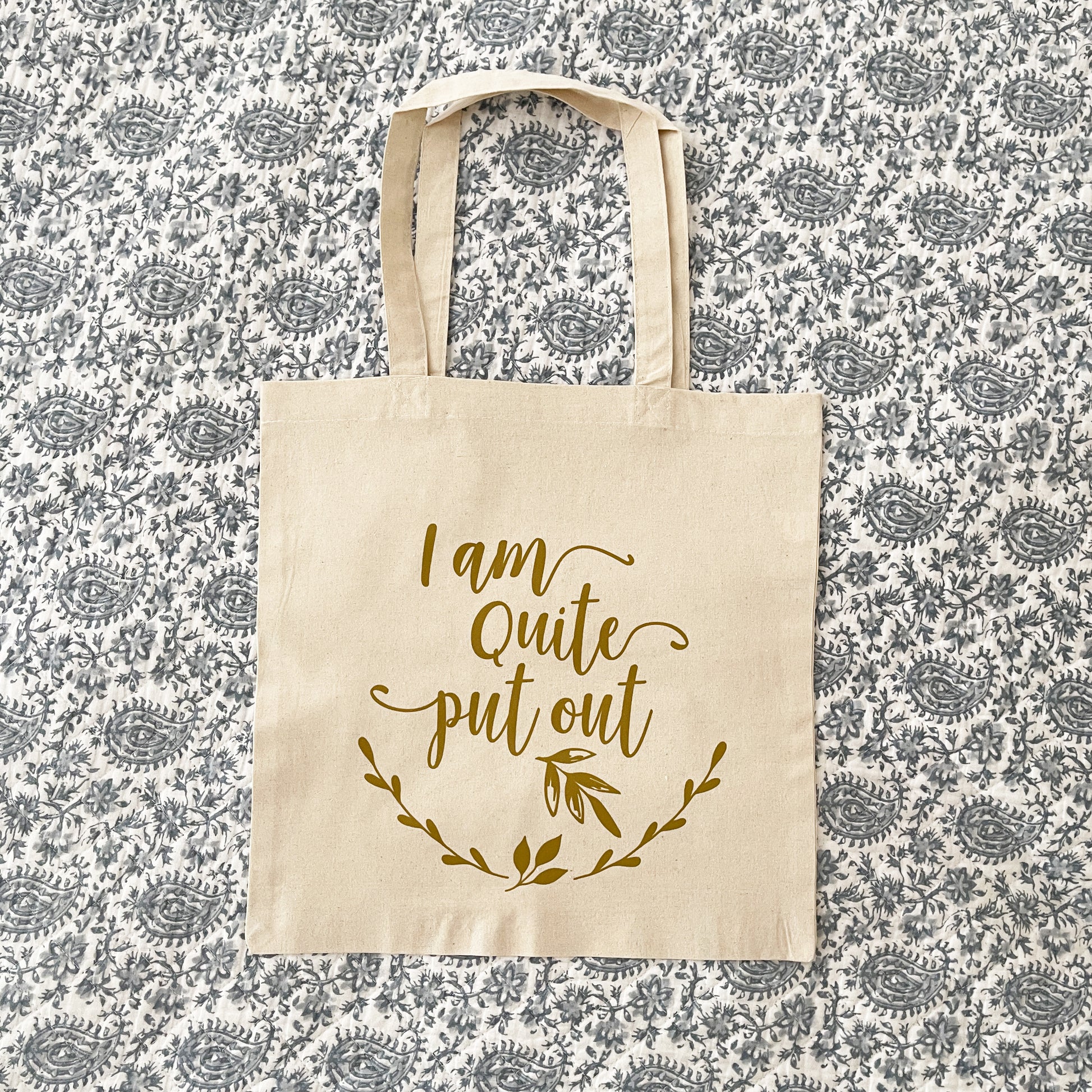 Natural cotton tote bag laying flat on paisley background, featuring the phrase 'I am quite put out' in elegant script with a leafy accent below. The text and design are printed in a soft, gold color, creating a minimalist and playful aesthetic.