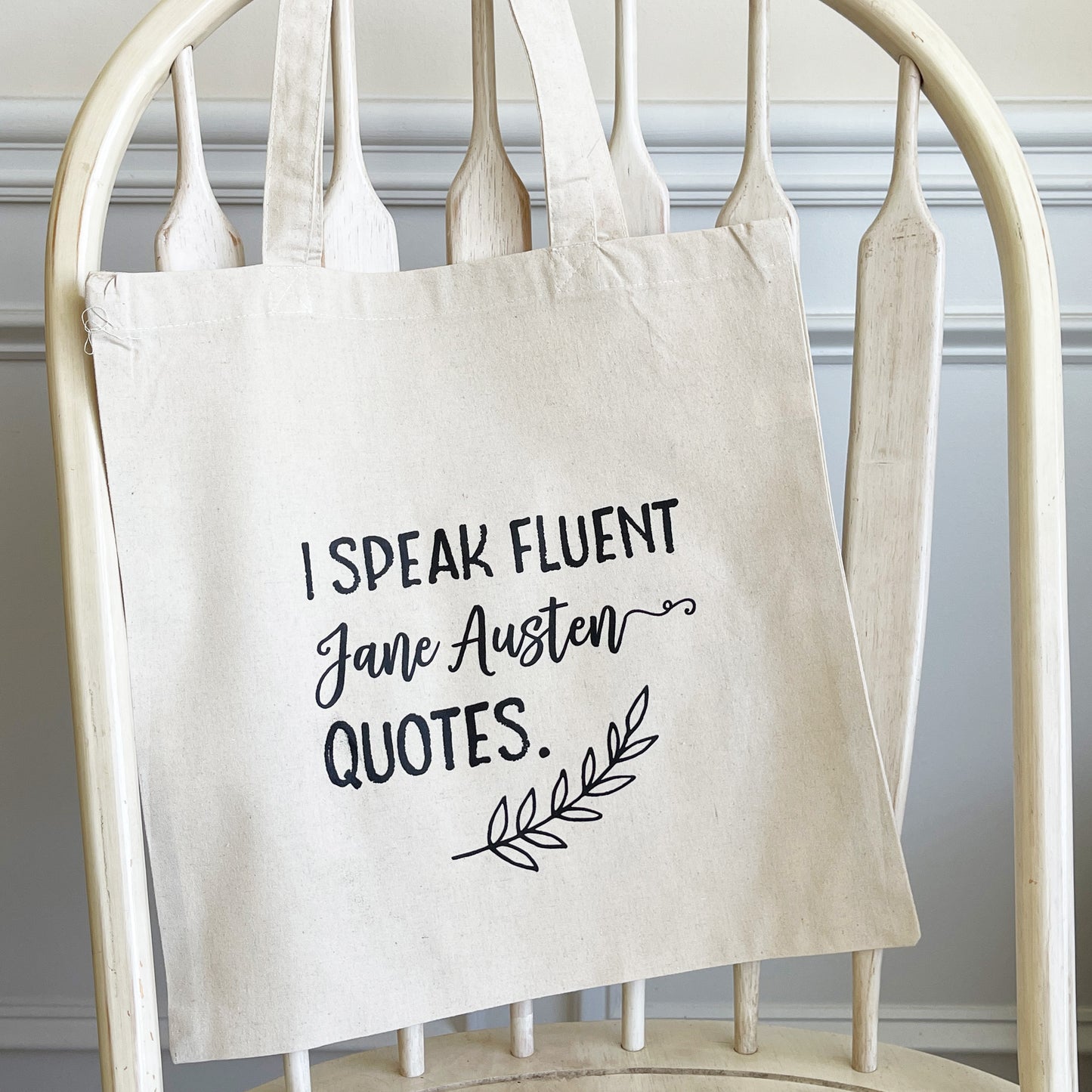 Natural cotton tote bag hanging on a white wooden chair, featuring the phrase 'I speak fluent Jane Austen quotes.' in bold black script, with a decorative leaf accent below. The tote has a minimalist and literary-inspired design, perfect for book lovers and Austen fans.