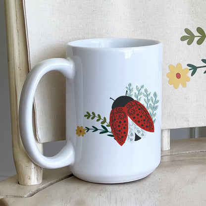 White mug featuring fa colorful illustration of a red ladybug with black spots, delicate floral accents, and green leaves near the bottom corner.