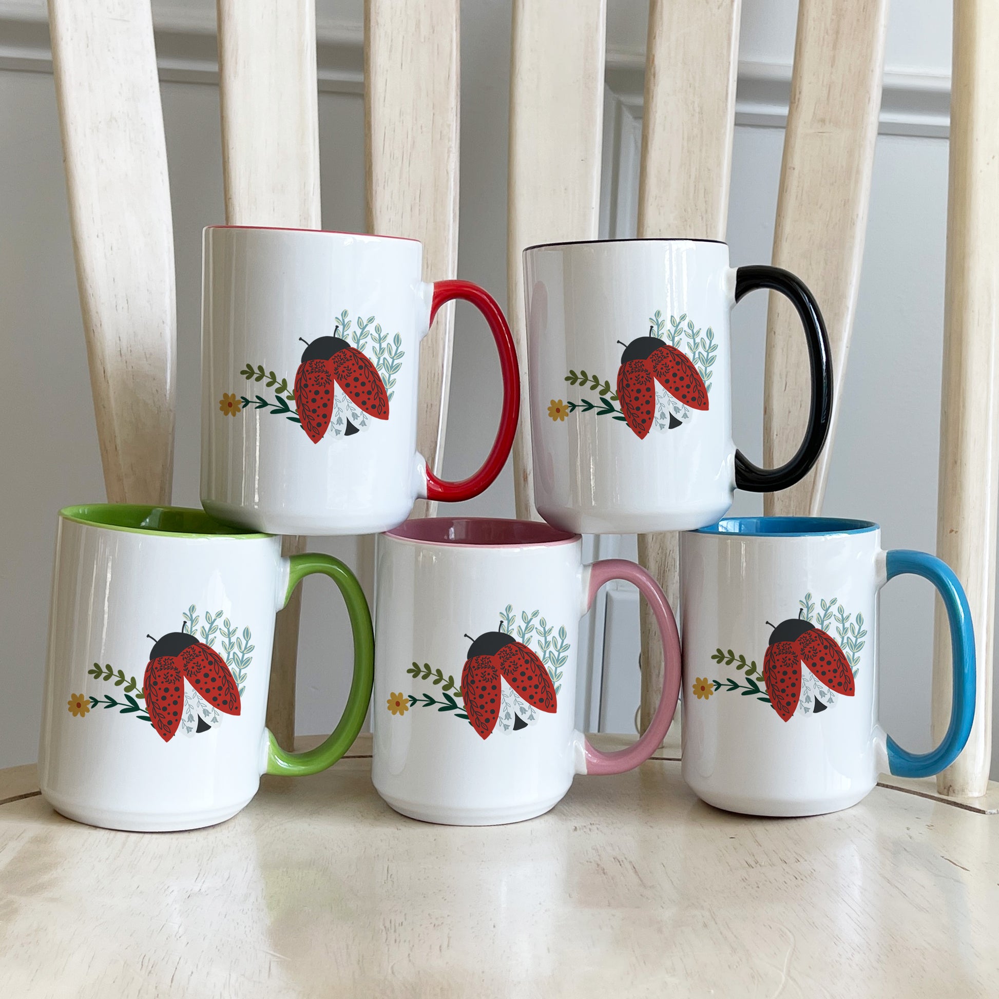 Five mugs with a colorful illustration of a red ladybug with black spots, delicate floral accents, and green leaves on each of the color accent mugs. The accent colors are red, black, green, pink, and blue.