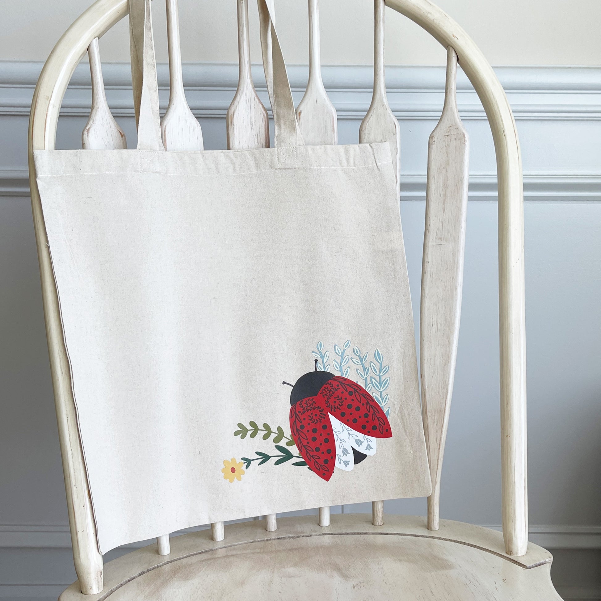 Beige canvas tote bag hanging on a white wooden chair, featuring a colorful illustration of a red ladybug with black spots, delicate floral accents, and green leaves near the bottom corner.