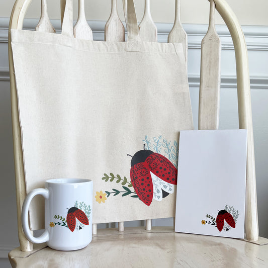 Coordinating gift set that features colorful illustration of a red ladybug with black spots, delicate floral accents, and green leaves canvas tote bag, notepads, and mug displayed on light wood chair.